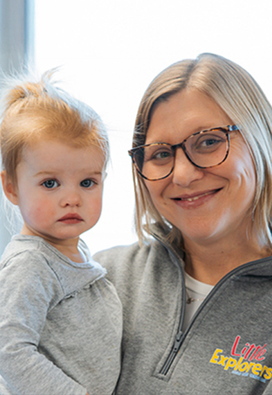 A smiling woman wearing glasses holds a young child with light brown hair. Both are dressed in light grey tops, and the woman’s shirt has a “Little Explorers” logo. They are indoors with natural light in the background.