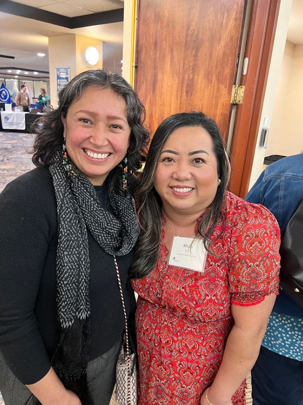 Two women smiling and standing close together indoors. One wears a black scarf and gray sweater, the other wears a red patterned dress with a conference name tag. A wooden door and people in the background are visible.