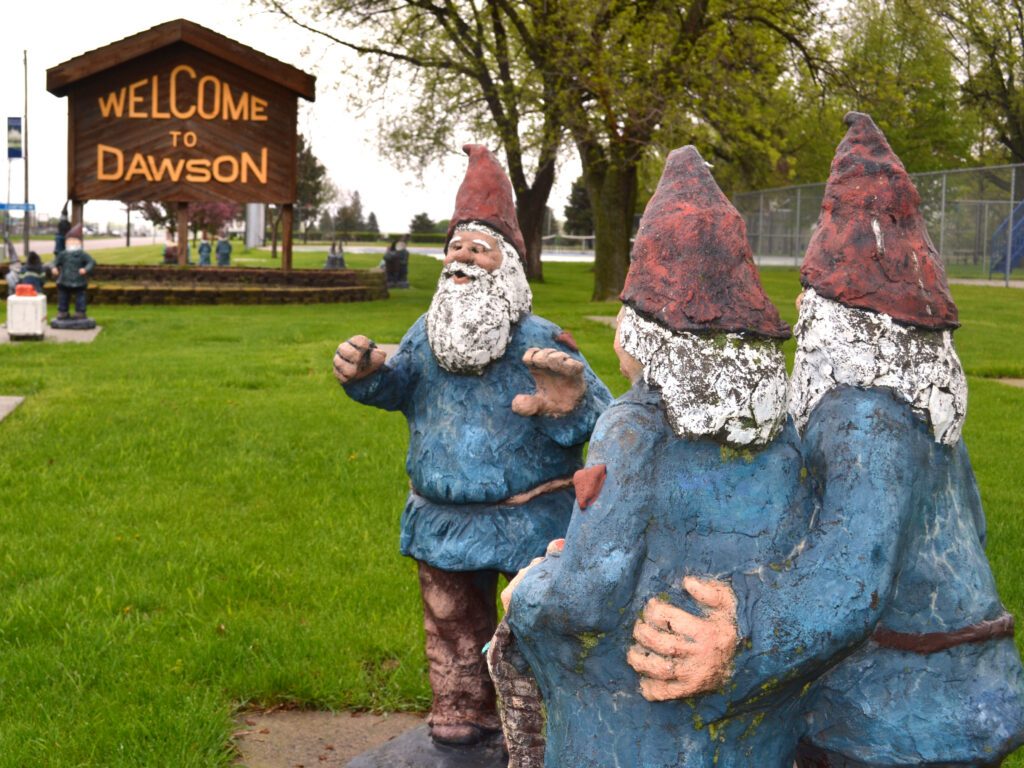 Three garden gnome statues stand near a grassy area, facing a large wooden “Welcome to Dawson” sign. Trees and additional gnome statues are visible in the background.