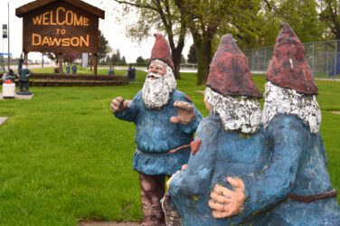 Three garden gnome statues stand near a grassy area, facing a large wooden “Welcome to Dawson” sign. Trees and additional gnome statues are visible in the background.