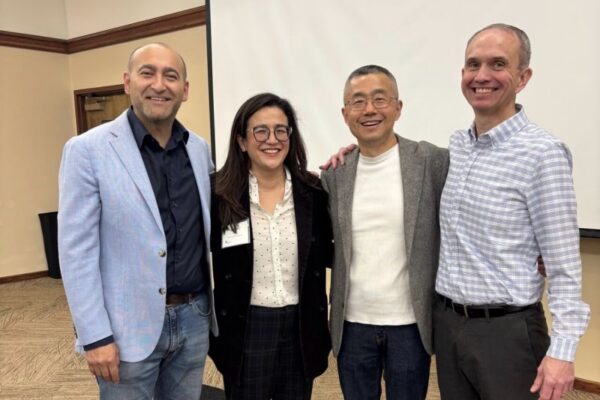 Four people stand together smiling in a conference room. They are dressed in business or business casual attire, with a projector screen and neutral walls in the background.