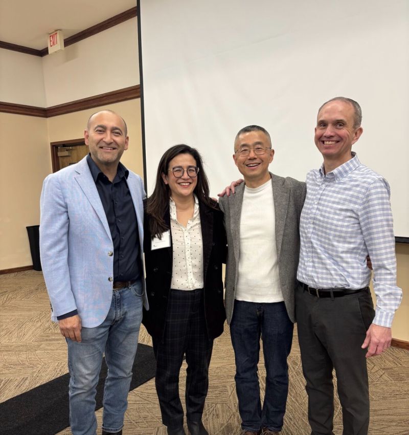 Four people stand together smiling in a conference room. They are dressed in business or business casual attire, with a projector screen and neutral walls in the background.