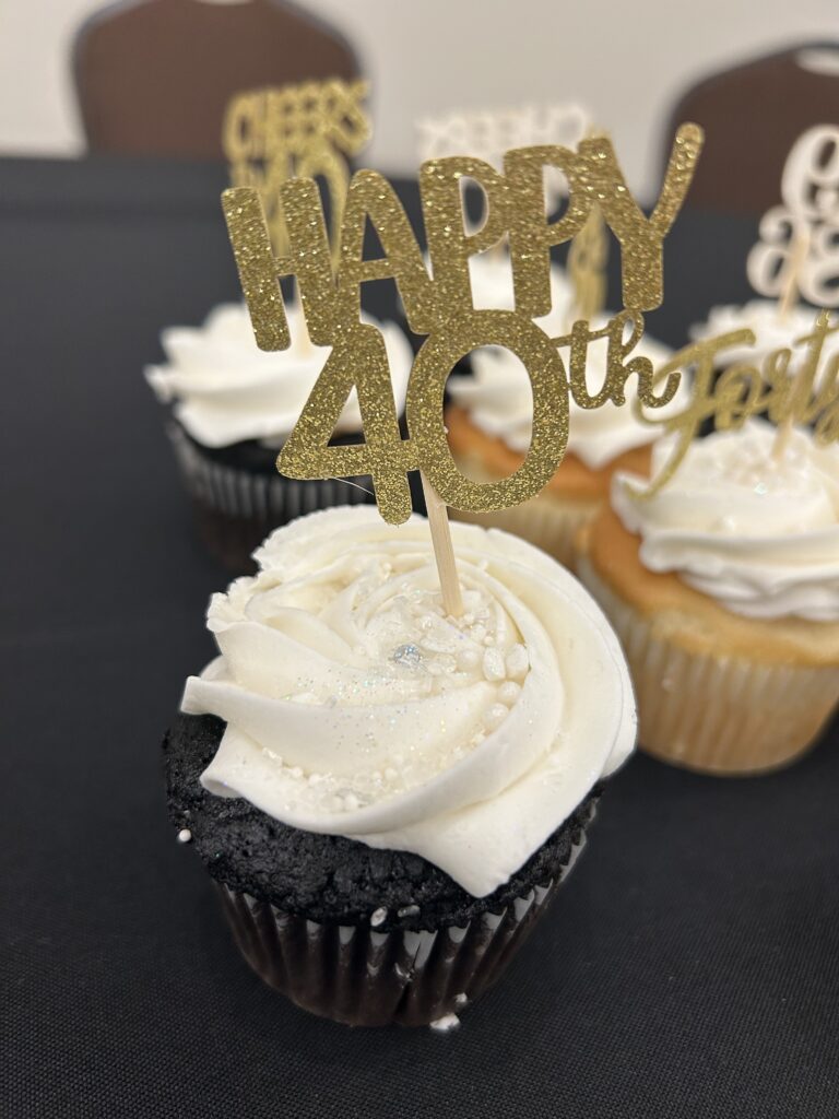 A chocolate cupcake with white frosting and glittery sprinkles has a gold HAPPY 40th topper. More cupcakes with similar toppers are in the background on a black tablecloth.