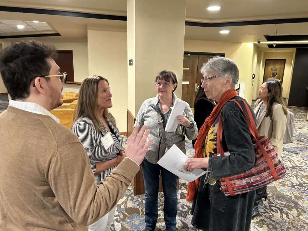 Five adults stand in a hotel lobby, engaged in conversation. Three hold papers or notebooks. One woman wears a colorful scarf and striped bag. The atmosphere appears friendly and professional.