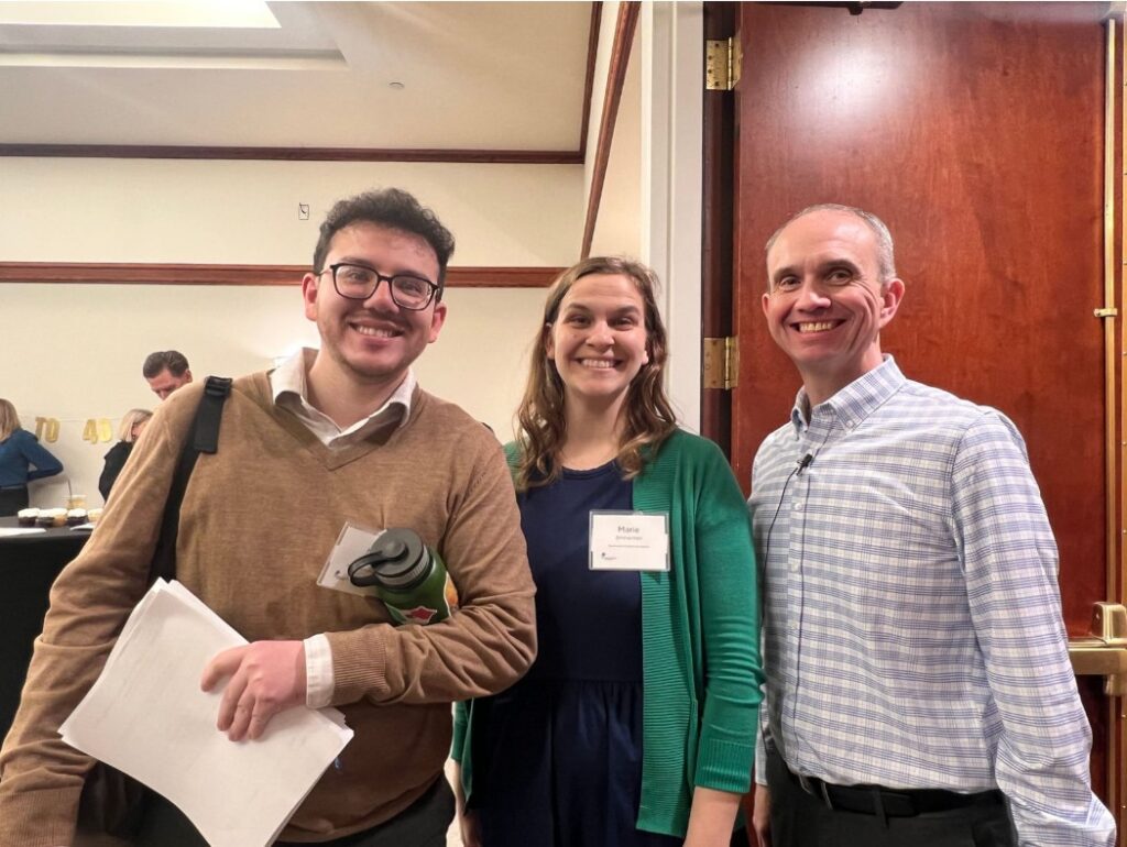 Three people stand indoors, smiling at the camera. The man on the left holds papers and a water bottle, while the woman and man beside him wear name tags. They are in a conference or event setting with wooden doors behind them.