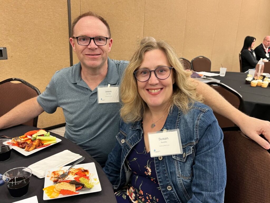 A man and woman, both wearing name tags, sit together at a round table with plates of food. They are smiling at the camera in a conference or banquet setting. Other people are visible in the background.