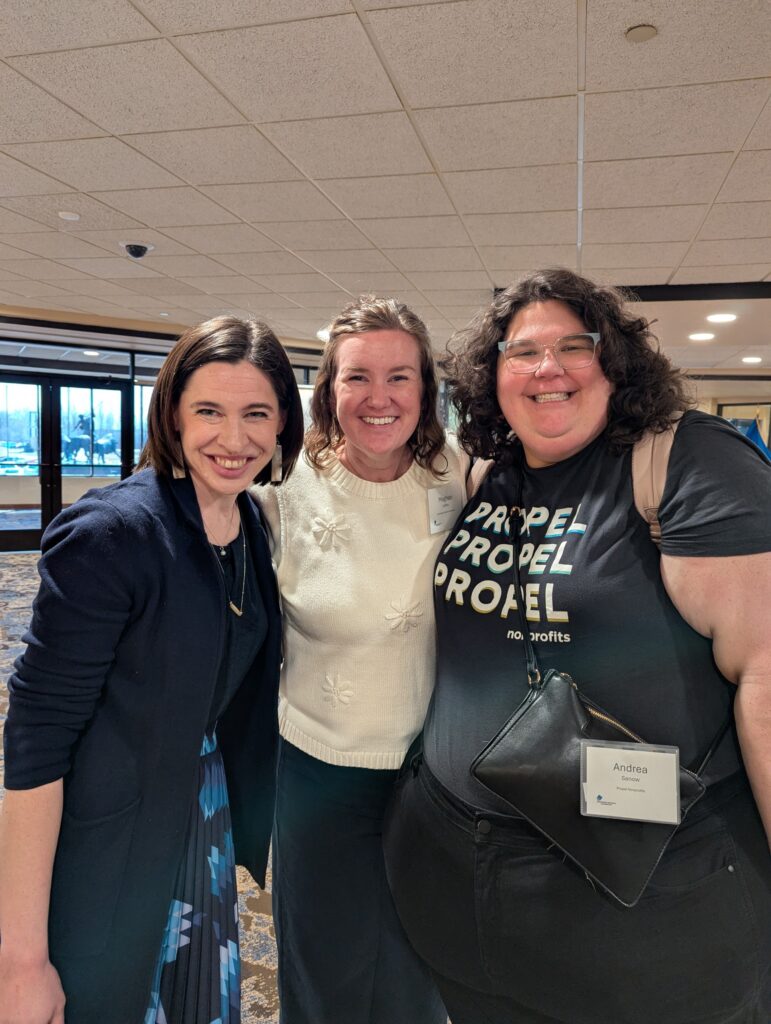 Three women smiling and posing together indoors at an event. One wears a black blazer, one a white sweater, and one a black t-shirt with PROPEL written on it, along with a name tag reading Andrea.