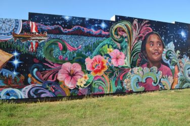 Colorful mural featuring a smiling girl, vibrant flowers, swirling waves, and a canoe with a red sail, set against a starry night sky.