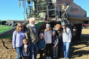 A group of seven people, including children and adults, stand smiling in front of a large Gleaner combine harvester in a sunlit cornfield with dry cornstalks on the ground. The sky is clear and blue.