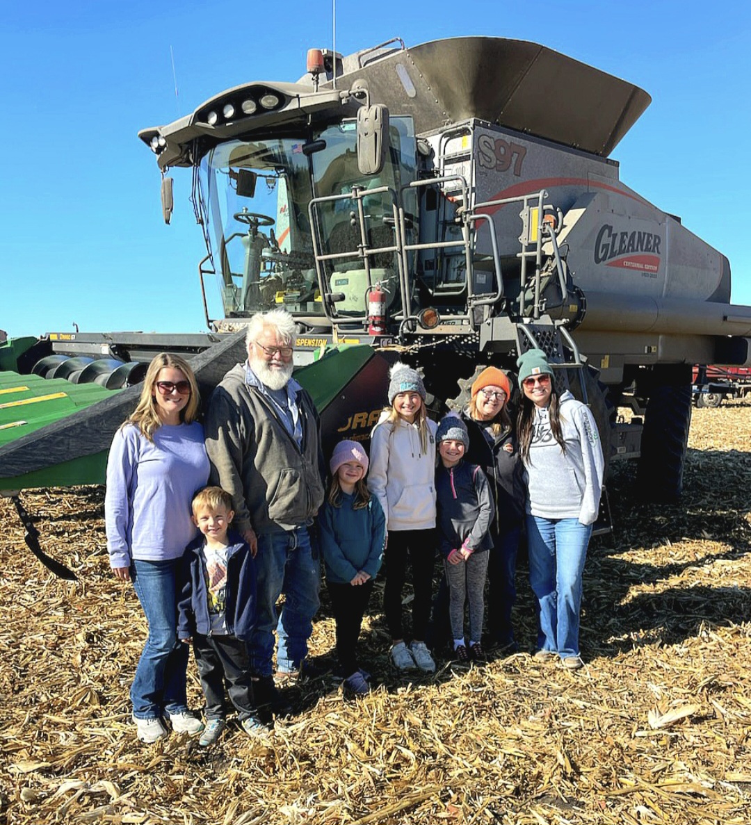A group of seven people, including children and adults, stand smiling in front of a large Gleaner combine harvester in a sunlit cornfield with dry cornstalks on the ground. The sky is clear and blue.