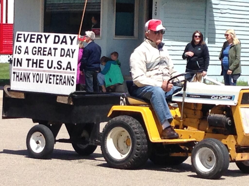 An older man wearing a red cap drives a yellow lawn tractor pulling a cart with a large sign that reads, EVERY DAY IS A GREAT DAY IN THE U.S.A. THANK YOU VETERANS. Several people stand in the background.