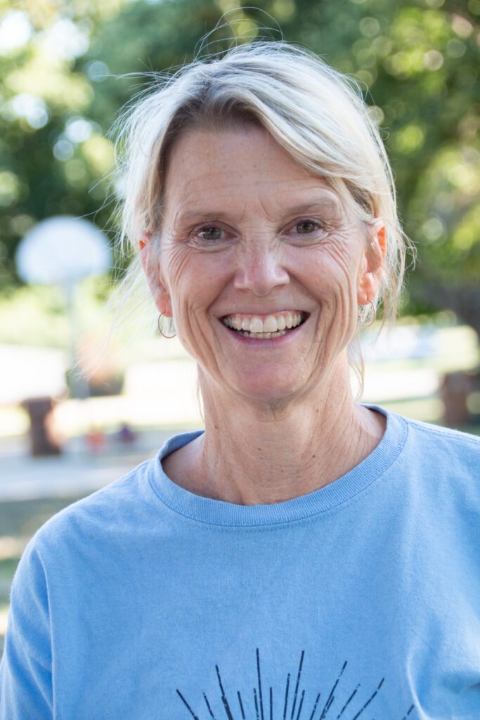 Ann smiles, her light blonde hair pulled back, wearing a light blue shirt and standing outdoors in a sunny park with green trees and a blurred basketball hoop in the background.