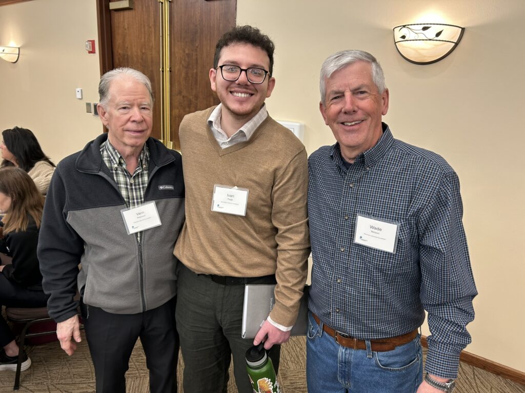 Three men stand smiling indoors at an event, each wearing name tags. The man on the left is in a black jacket, the middle man wears glasses and a tan sweater, and the man on the right is in a blue checked shirt.