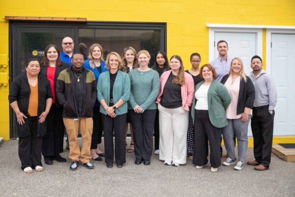 The Growing Local: Emerging Leaders cohort gathers for a group photo, smiling in front of a bright yellow building with white doors. They are posed outdoors on a gray pavement.