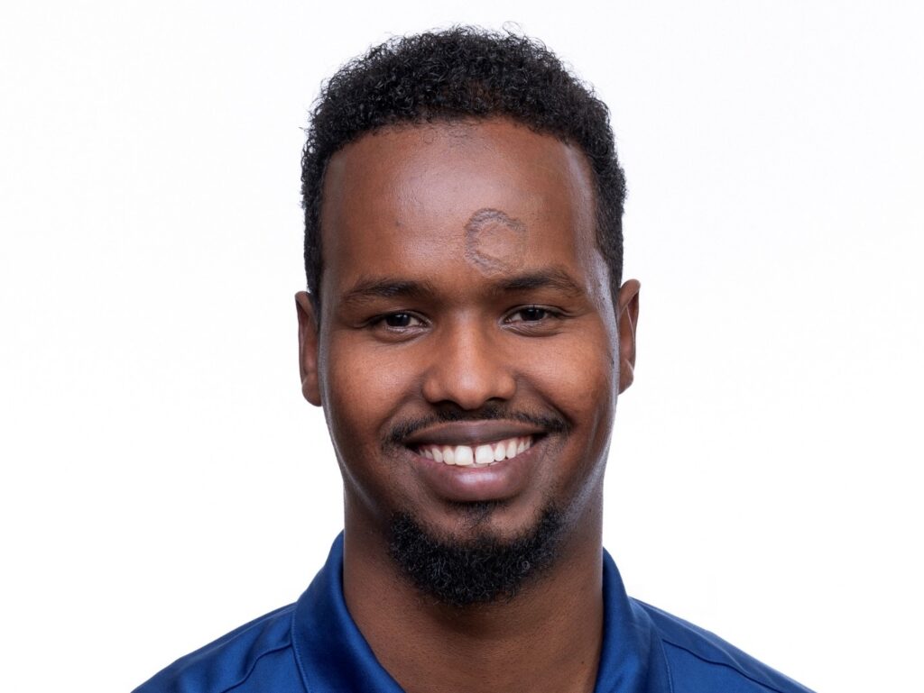 A man with short curly hair and a goatee smiles at the camera. He is wearing a blue polo shirt with the text Community Connector Services embroidered on it. The background is plain white.