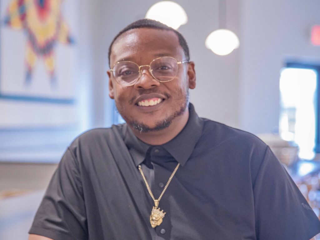 A man wearing glasses, a black shirt, gold jewelry, and beaded bracelets smiles while leaning on a counter in a modern, well-lit cafe with hanging lights in the background.