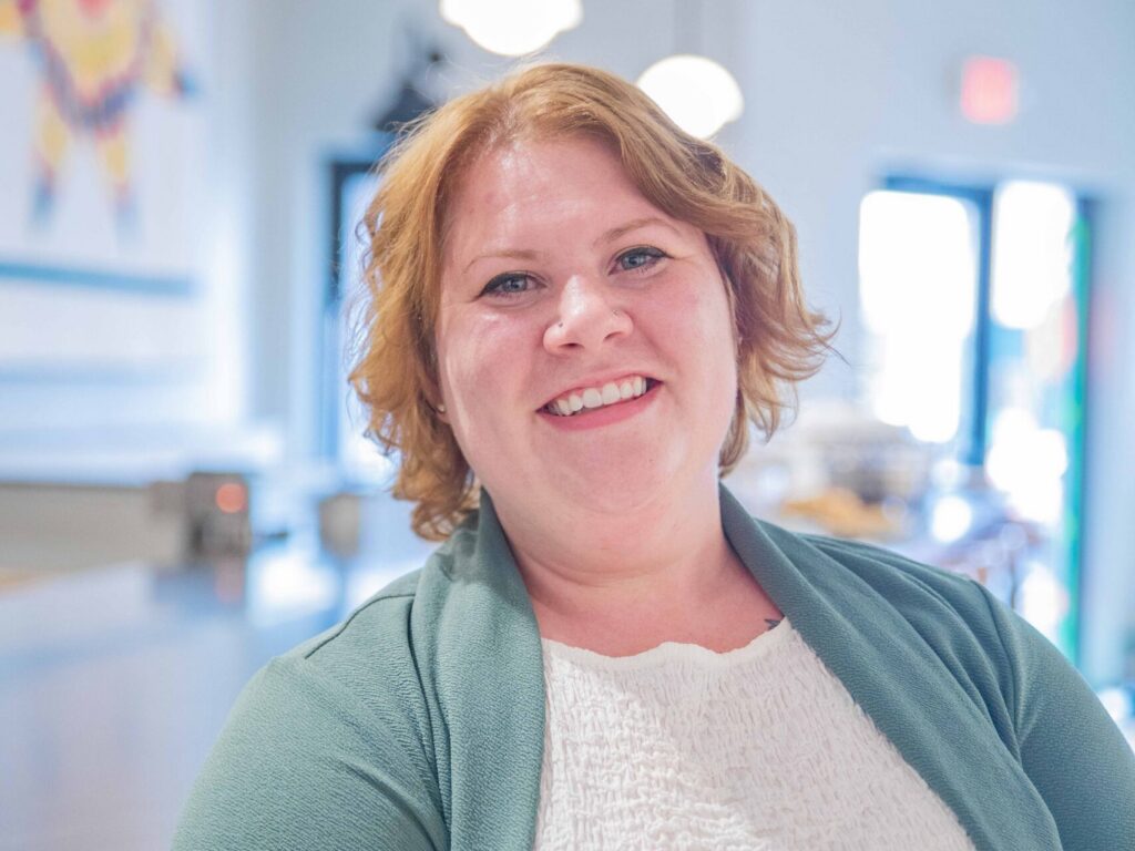 A woman with short, wavy red hair, wearing a light green cardigan and white top, smiles while leaning against a counter in a brightly lit, modern café with hanging lights.