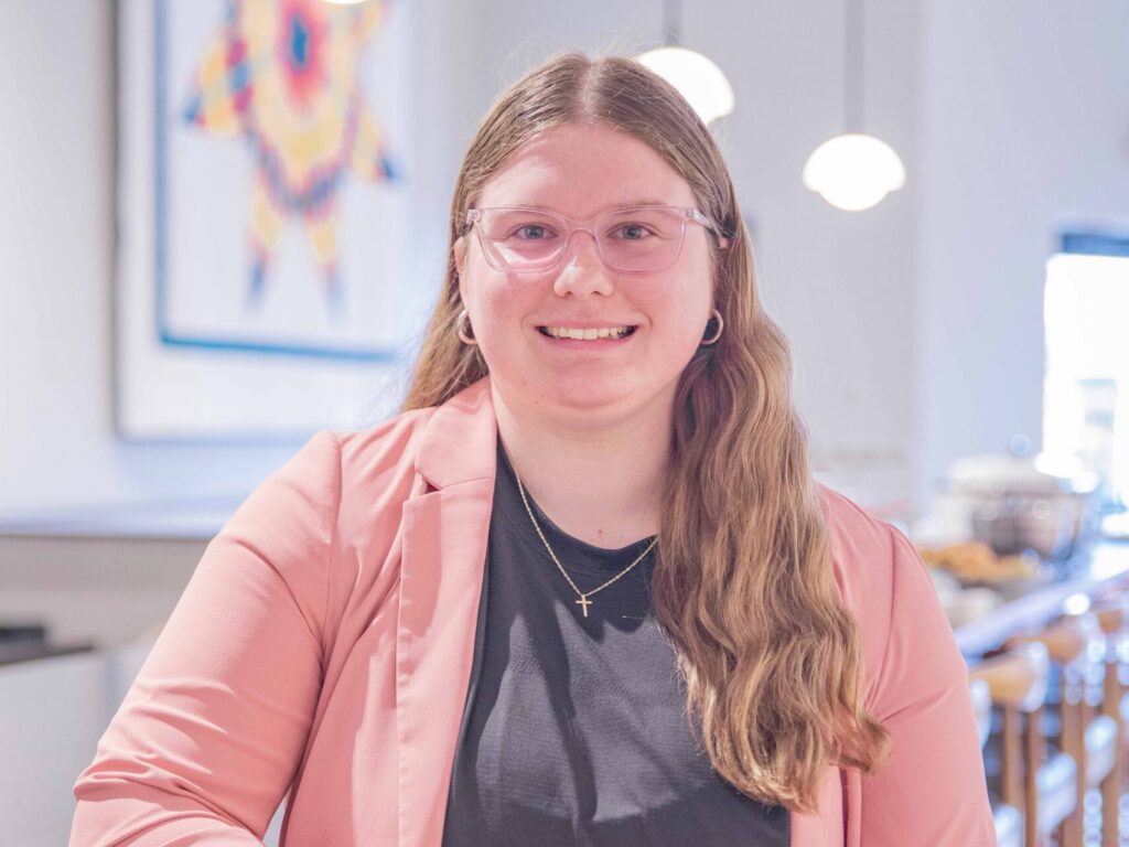 A person with long, light brown hair and glasses, wearing a pink blazer over a black shirt, stands indoors by a counter, smiling at the camera. Round lights and colorful artwork are visible in the background.