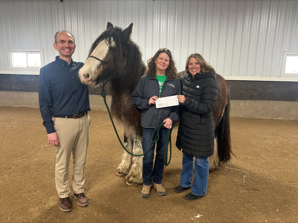 Three adults stand in a riding arena with a large horse. One woman holds a piece of paper, while the others smile at the camera. The setting is indoors with a dirt floor and white paneled walls.