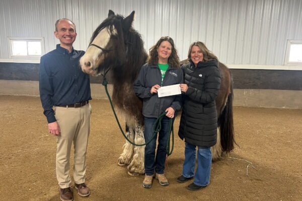 Three adults stand in a riding arena with a large horse. One woman holds a piece of paper, while the others smile at the camera. The setting is indoors with a dirt floor and white paneled walls.