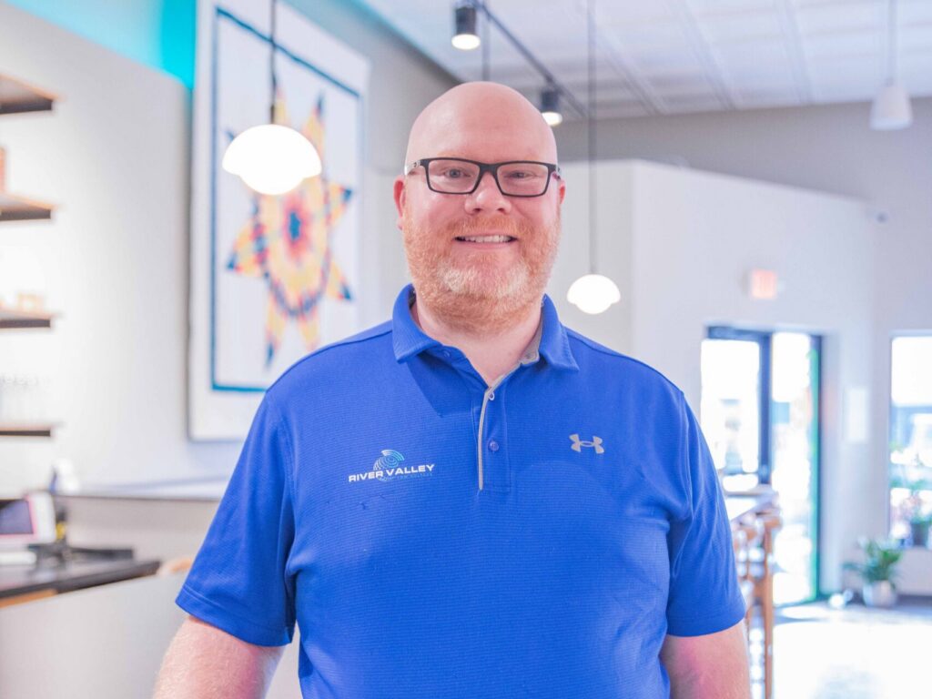 A smiling man in glasses and a bright blue polo shirt stands indoors by a counter in a modern, brightly lit room with patterned floor tiles and artwork on the wall.