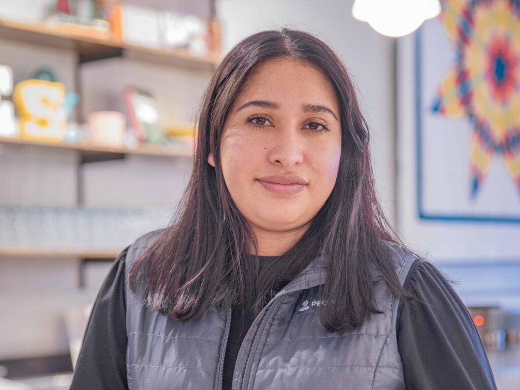A person with long dark hair wearing a gray vest and black shirt stands indoors, resting their hands on a counter with shelves and a colorful geometric artwork in the background.