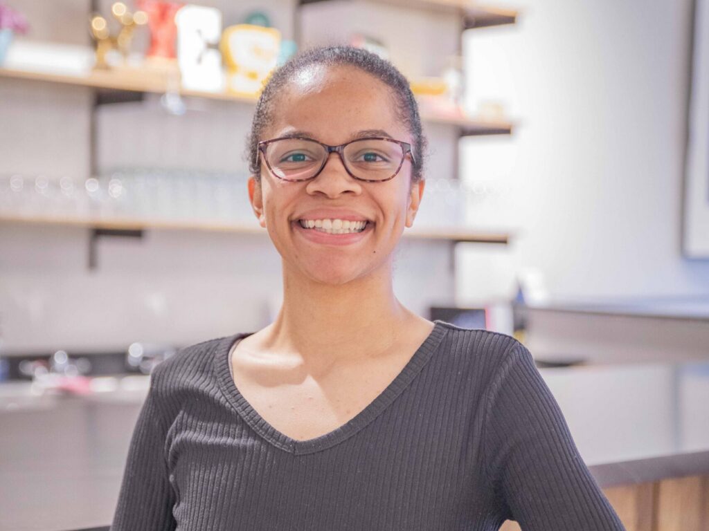 A woman with glasses and a black sweater smiles while sitting on a wooden chair in a modern kitchen with open shelves and a countertop in the background.