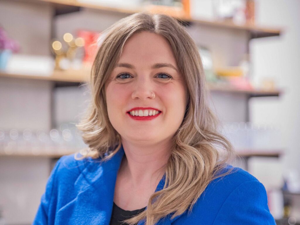 A woman with wavy blonde hair wearing a bright blue blazer and black top smiles, standing indoors with shelves and various items blurred in the background.