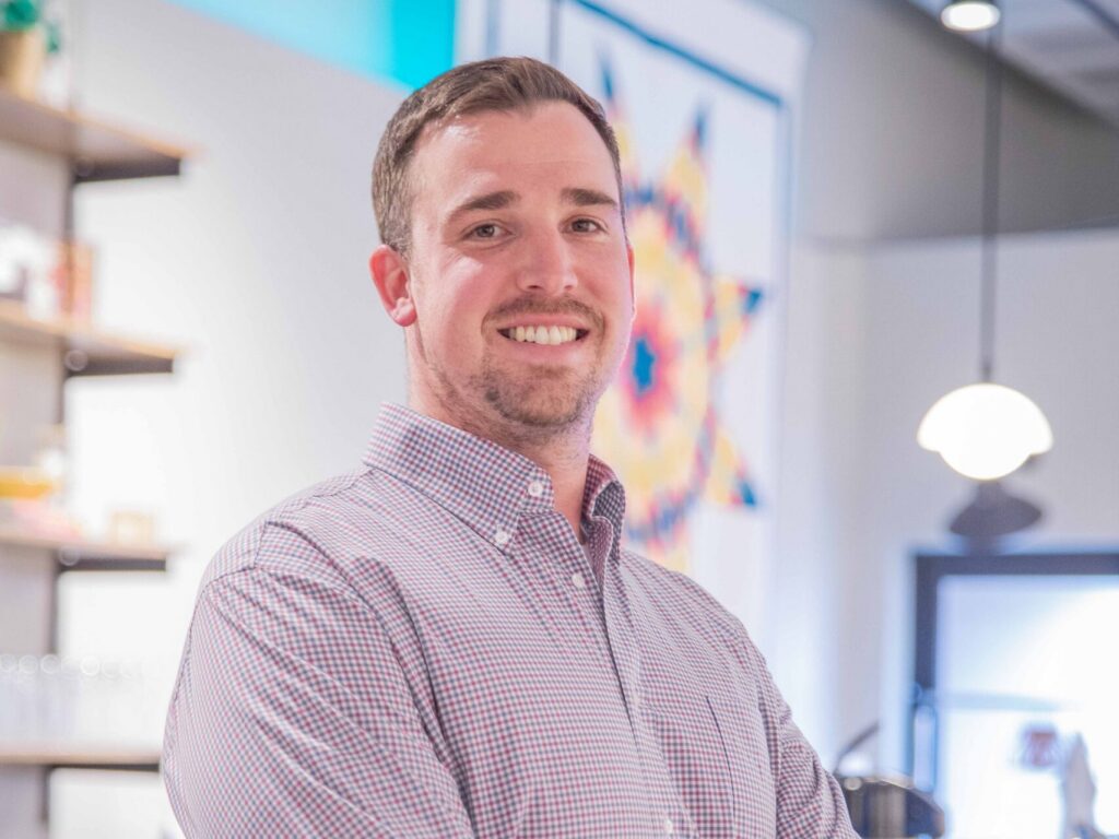 A man in a checked dress shirt stands with arms crossed, smiling, in a modern, brightly lit café with shelves and colorful artwork in the background.
