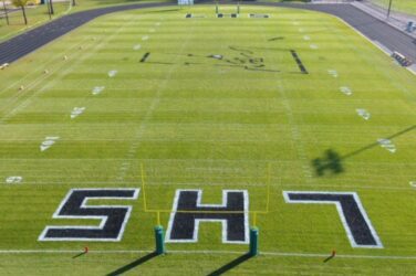 A football field with bright green turf, white yard lines, and large black and white SH7 letters painted in each end zone. The field is empty and seen from a high vantage point.