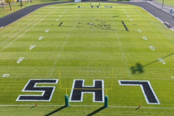 A football field with bright green turf, white yard lines, and large black and white SH7 letters painted in each end zone. The field is empty and seen from a high vantage point.