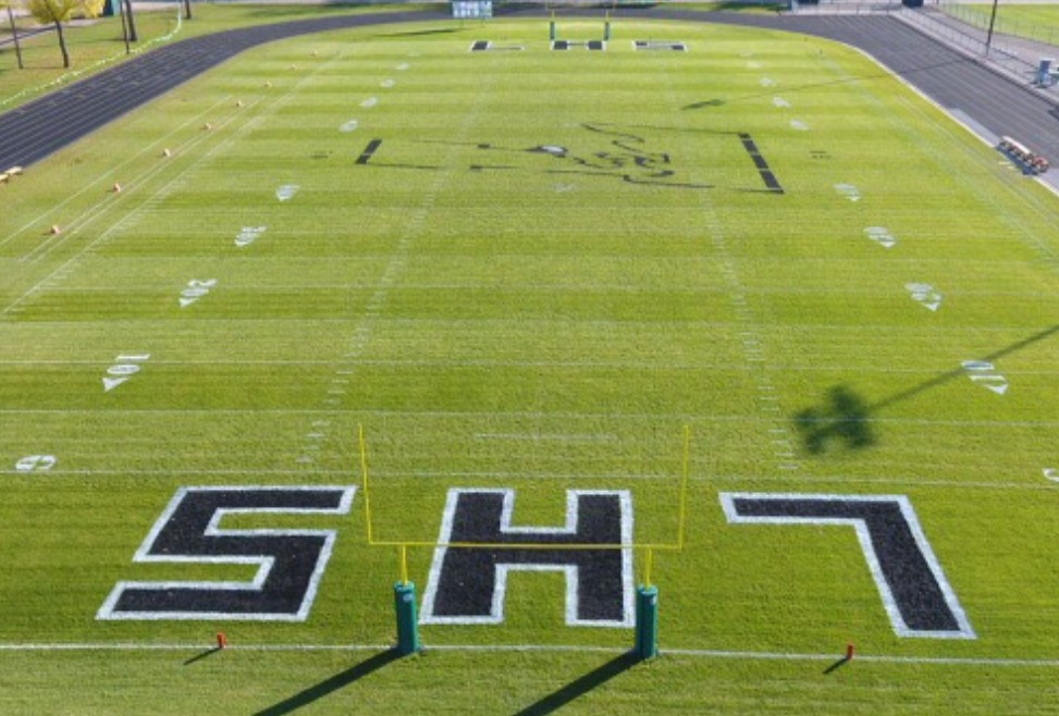 A football field with bright green turf, white yard lines, and large black and white LHS letters painted in each end zone.