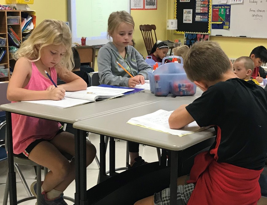 Three young children sit at a classroom table, focused on writing in notebooks. The classroom has colorful decorations, a whiteboard, and other students working in the background.