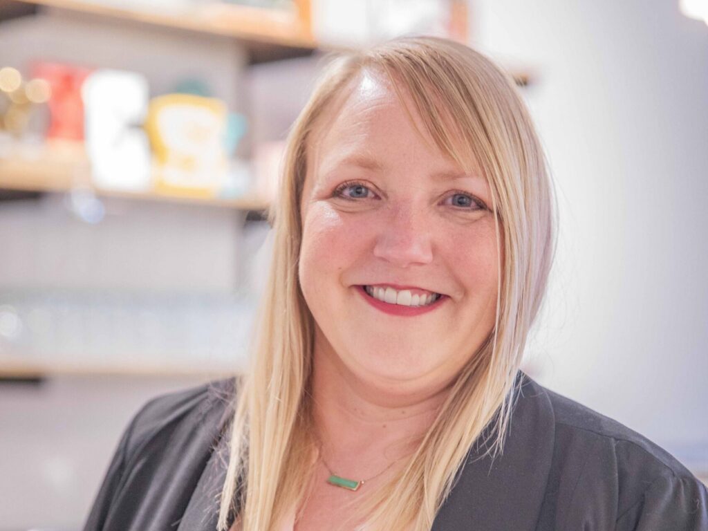 A woman with straight blonde hair and a black blazer over a light pink blouse smiles while standing in a modern kitchen with shelves and dishes in the background.
