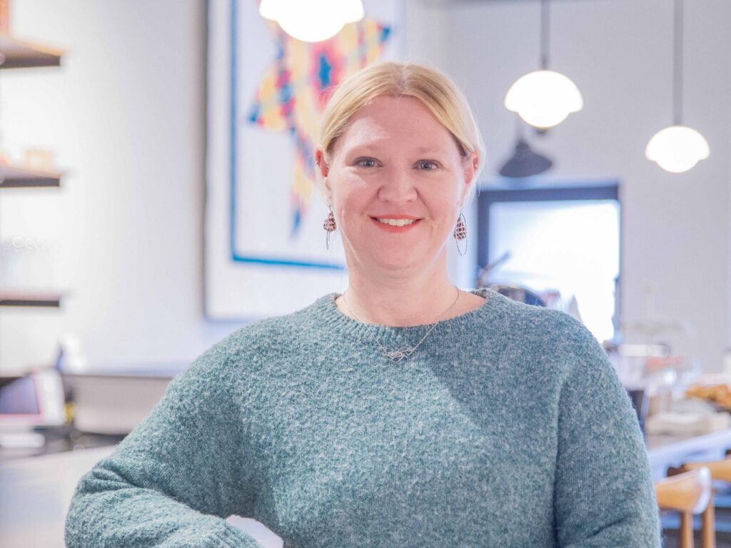A woman with blonde hair, wearing a teal sweater, stands smiling and leaning against a wooden counter in a modern, brightly lit cafe with pendant lights and a colorful quilt on the wall behind her.