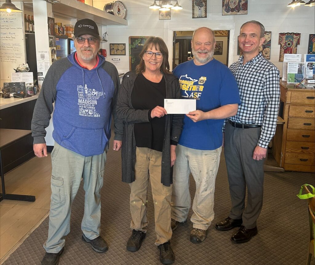 Four adults stand indoors, smiling, with one woman handing a check to the man beside her. Two men wear casual clothes and hats, one woman wears glasses, and one man is dressed in business attire. The setting appears to be a cozy café.