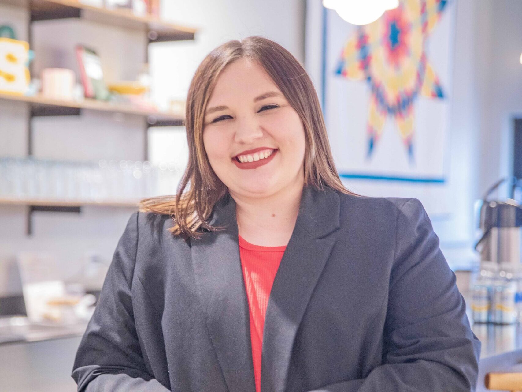 A woman with straight brown hair, wearing a black blazer and red top, smiles while leaning against a kitchen counter. Bright shelves and a colorful geometric wall hanging are visible in the background.