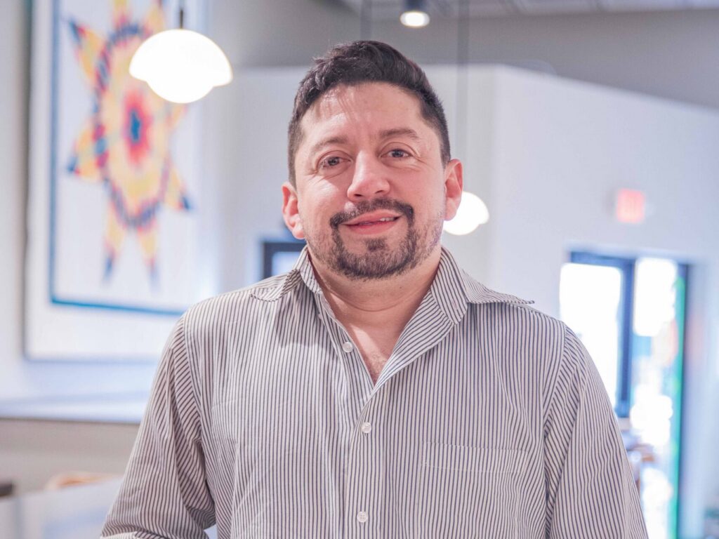 A man with short dark hair and a trimmed beard, wearing a striped button-up shirt and black pants, stands indoors, smiling, with one hand resting on a counter. Modern decor and a bright quilted artwork are visible in the background.