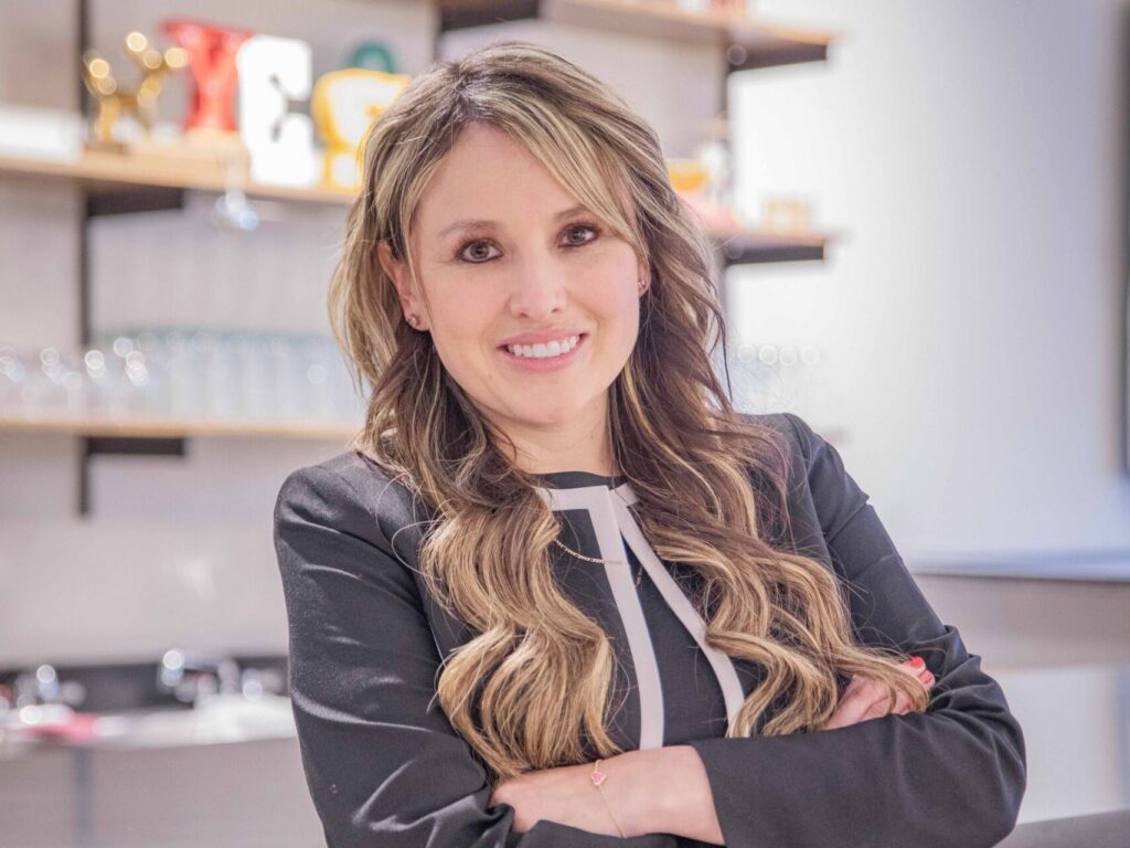 A woman with long wavy hair, wearing a black suit with white accents, stands smiling with her arms crossed in front of a bar or counter with shelves and glasses in the background.