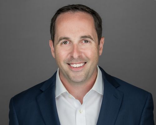 Smiling man with short dark hair wearing a white dress shirt and dark blue blazer, posing in front of a plain gray background.
