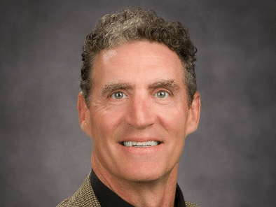 A middle-aged man with short, curly brown hair is smiling while wearing a brown checked blazer and a black collared shirt, posed in front of a plain, dark gray background.