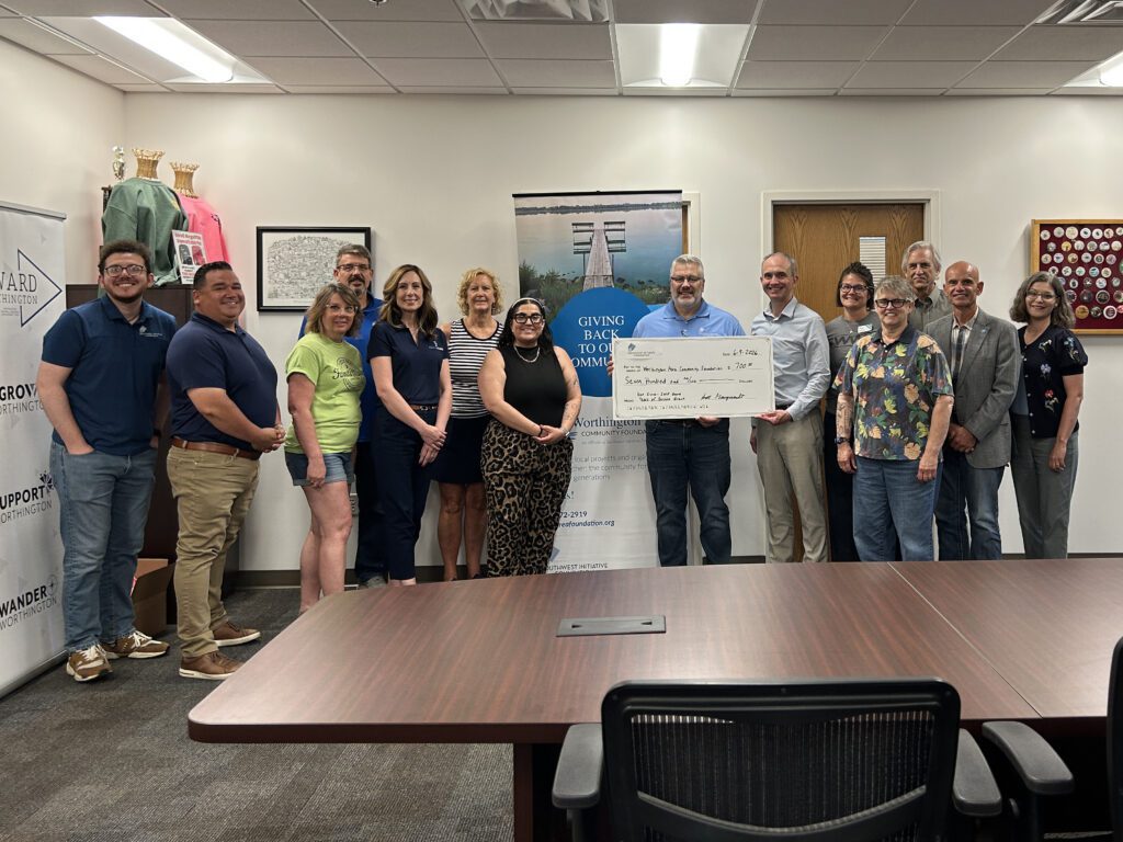 Fifteen people stand in a conference room, smiling at the camera. A group in the center holds a large ceremonial check. Banners, trophies, and framed pictures are visible in the background.