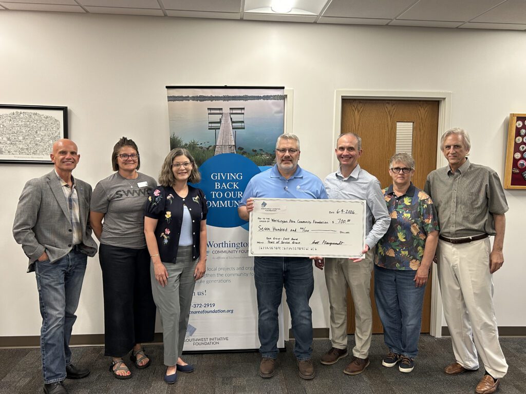 Seven people stand in a row smiling, as two of them hold a large ceremonial check in front of a banner that reads Giving Back To Our Community in an office setting.