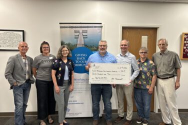 Seven people stand in a row, some smiling, as two of them hold a large ceremonial check in front of a banner that reads Giving Back To Our Community in an office setting.