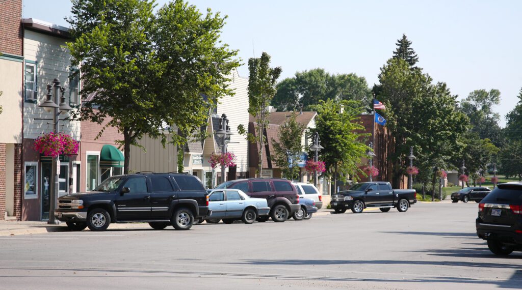 A small town main street with parked cars, storefronts, hanging flower baskets, trees, and American flags on a sunny day.