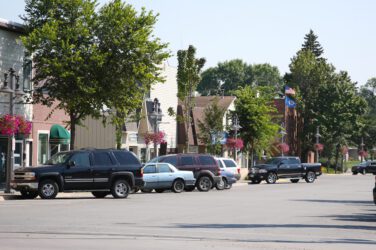 A small town main street with parked cars, storefronts, hanging flower baskets, trees, and American flags on a sunny day.