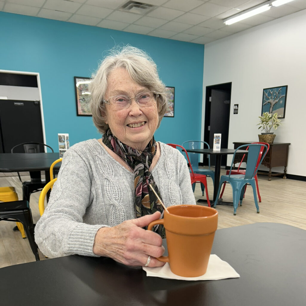 Smiling elderly woman with gray hair, glasses, and a scarf sits at a table in a brightly lit cafe, holding a terracotta mug placed on a napkin.