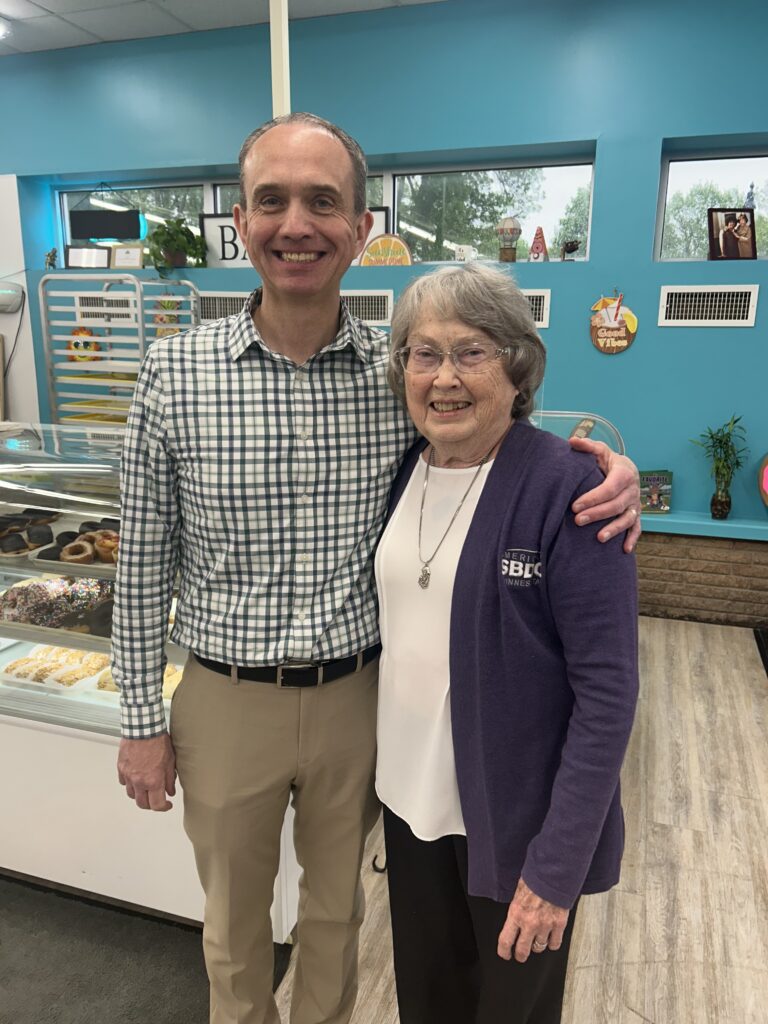 A smiling man in a checkered shirt stands next to an older woman in a purple cardigan inside a bakery. Pastries are visible in a display case behind them, and the walls are painted bright blue.