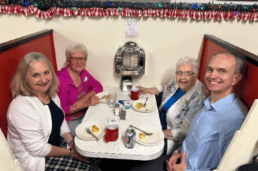 Four people sit in a retro diner booth, smiling at the camera. There are plates with food, mugs, and a jukebox on the wall behind them. The booth is decorated with festive garland.
