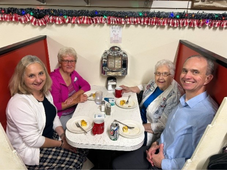 Four people sit in a retro diner booth, smiling at the camera. There are plates with food, mugs, and a jukebox on the wall behind them. The booth is decorated with festive garland.